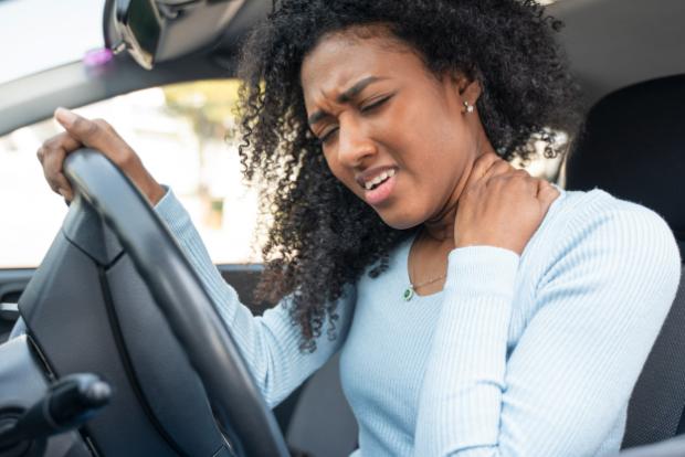 Attractive black woman sits behind steering wheel of car with one hand cradling the back of her neck as she winces in pain.