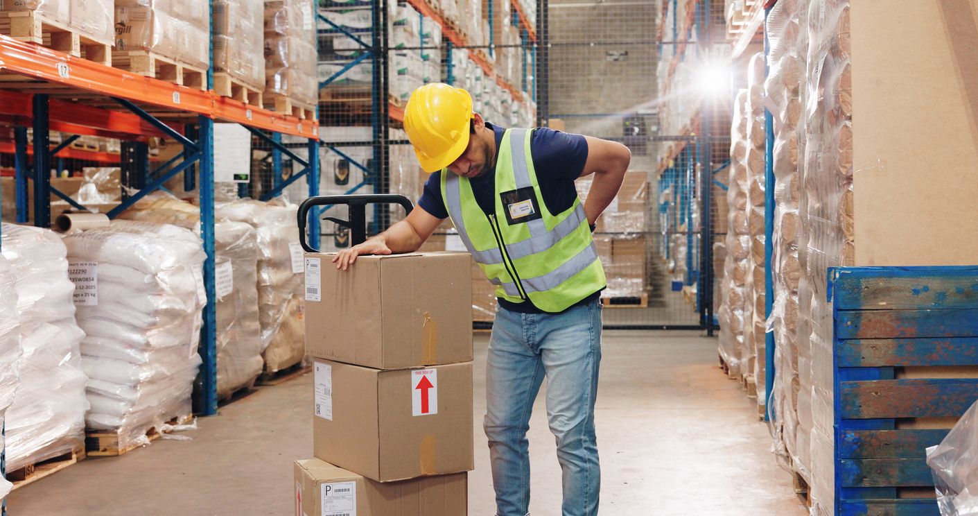 Warehouse worker wearing a yellow hard hat and reflective vest leaning on a stack of boxes and rubbing his aching back with his left hand.