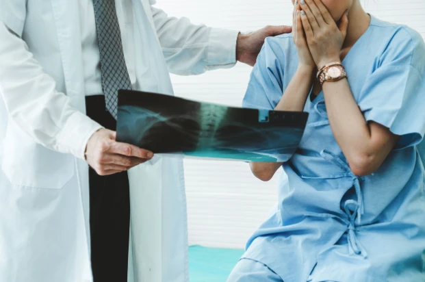 Doctor holds X-ray to patient in hospital room as patient covers mouth with hands in shock